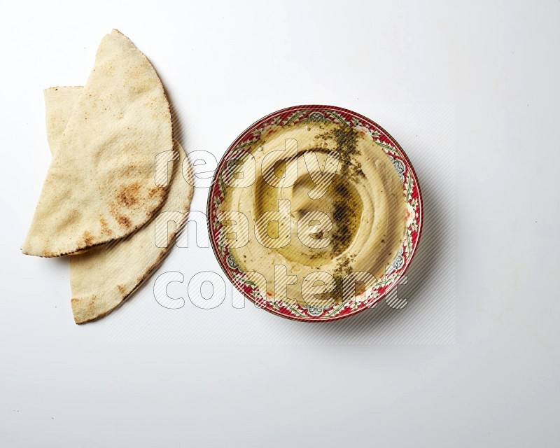 Hummus in a red plate with patterns garnished with zaatar & sumak on a white background