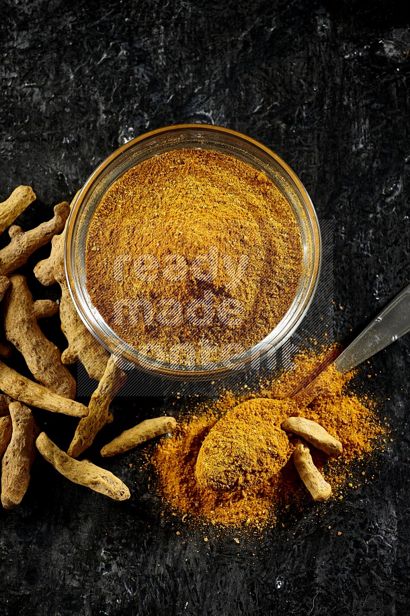 A glass bowl and a metal spoon full of turmeric powder and dried whole fingers next of them on textured black flooring