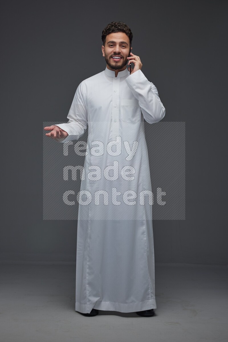 A Saudi man wearing Thobe and talking in the phone while standing and making different poses eye level on a gray background