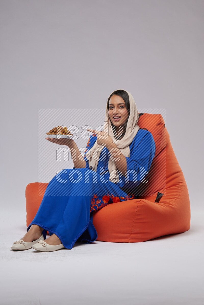 A Woman sitting on an orange beanbag wearing Jalabeya holding a plate of dates