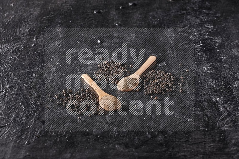 2 wooden spoons full of black pepper powder and black pepper beads spread on a textured black flooring
