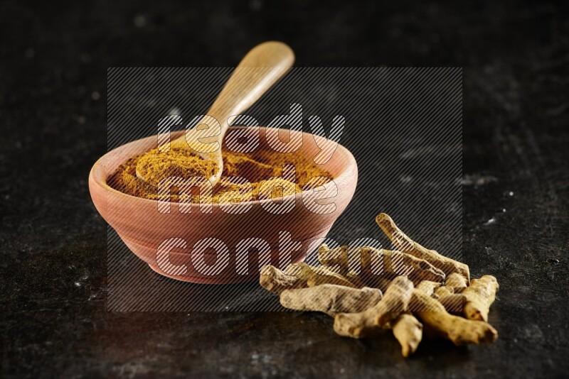 A wooden bowl and a wooden spoon full of turmeric powder with dried turmeric fingers on textured black flooring