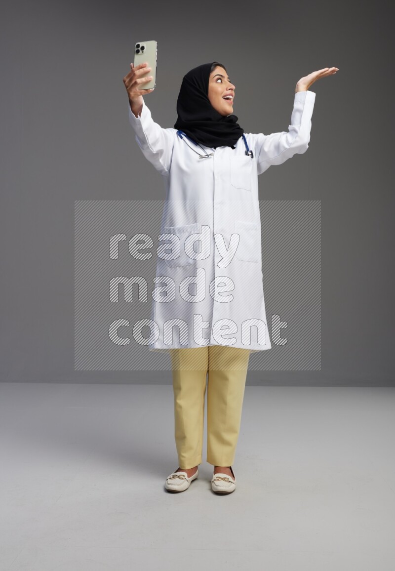 Saudi woman wearing lab coat with stethoscope standing taking selfie on Gray background