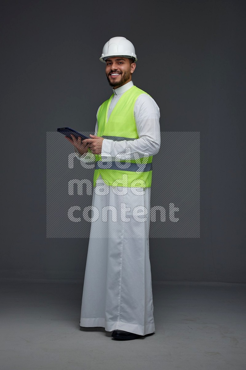 A Saudi man wearing Thobe with a yellow safety vest and white helmet standing and using his tablet different angles eye level on a gray background