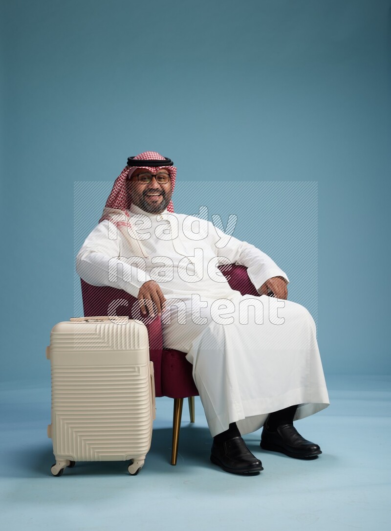 Saudi Man with shimag sitting on chair with a travel luggage on blue background