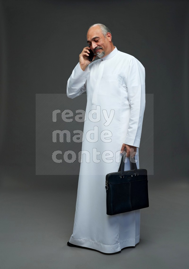 Saudi man without shomag Standing holding bag and talking on phone on gray background