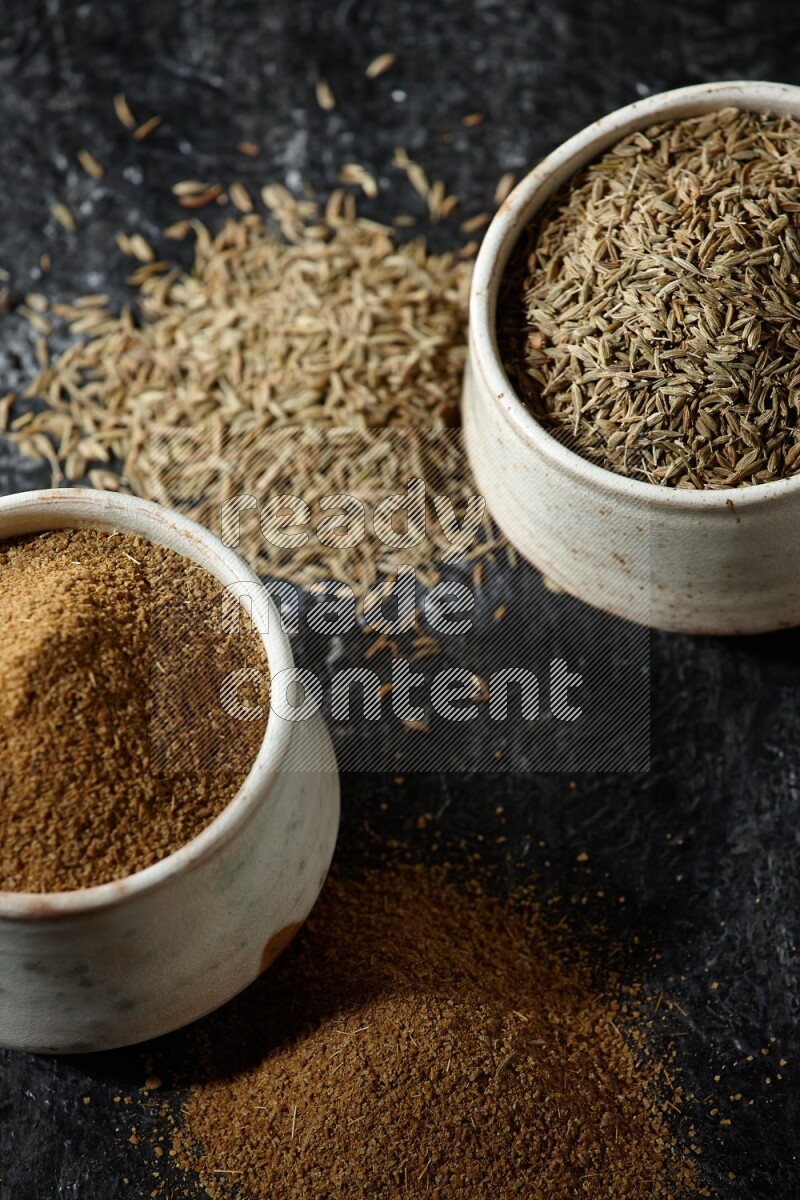 وعاءين بيج، أحدهما ممتلئ ببودرة الكمون والآخر ممتلئ بحبوب الكمون، على خلفية سوداء