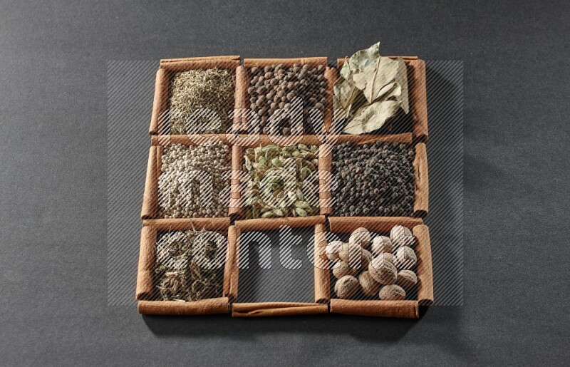 9 مربعات من أعواد القرفة ممتلئة بالهيل في المنتصف محاطة بجوزة الطيب والفلفل الأسود وورق الغار والبهارات والكمون والفلفل الأبيض والريحان المجفف والملح على أرضية سوداء