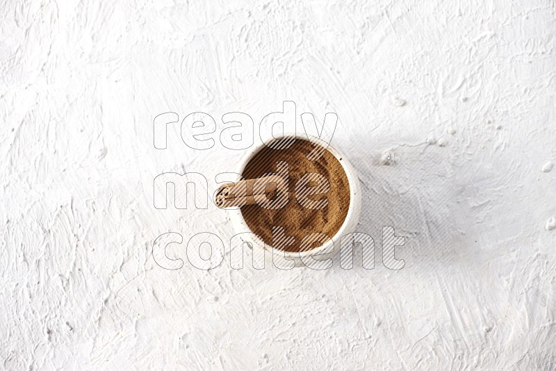 Ceramic beige bowl full of cinnamon powder with a cinnamon stick on a textured white background