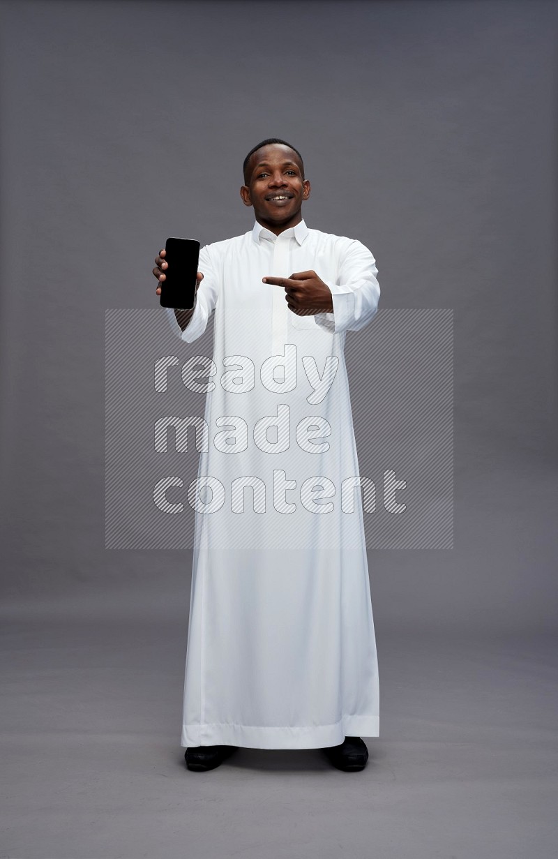 Saudi man wearing thob standing showing phone to camera on gray background