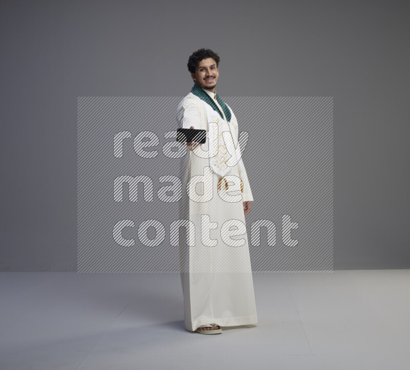 A Saudi man standing wearing thob and Saudi flag scarf showing phone to camera on gray background