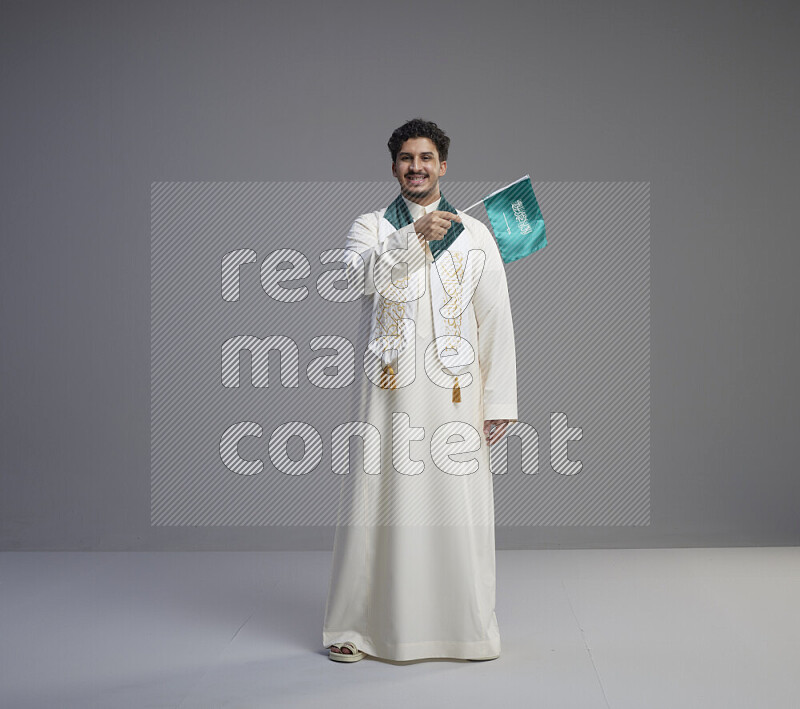 A Saudi man standing wearing thob and saudi flag scarf and holding small saudi flag on gray background