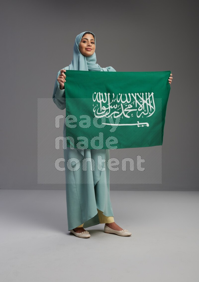 Saudi Woman wearing Abaya standing holding Saudi flag on Gray background
