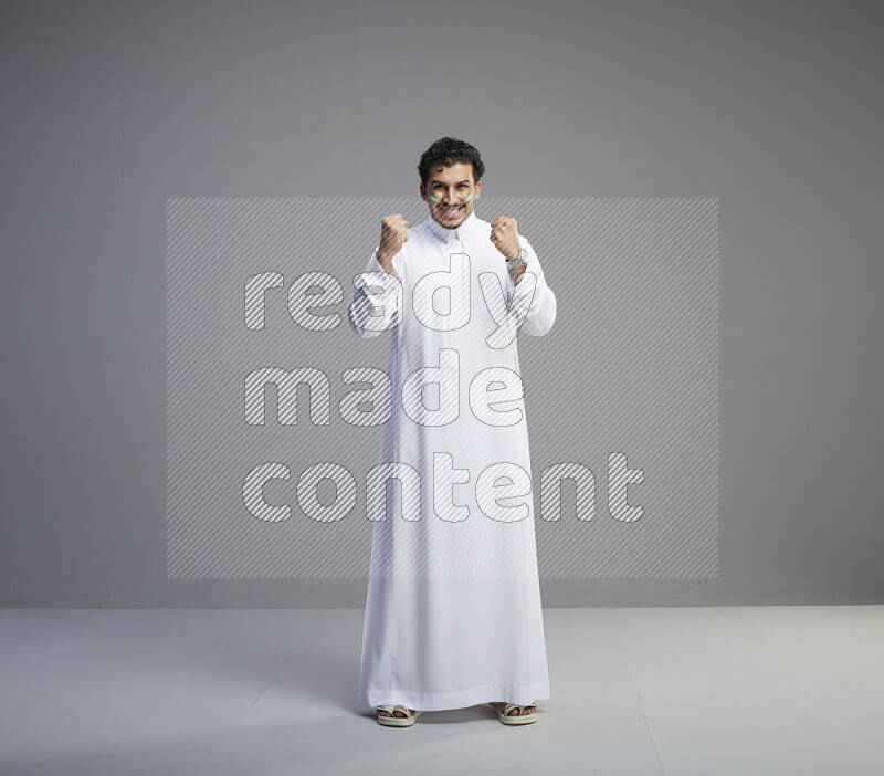A Saudi man standing wearing thob with face painting interacting with the camera on gray background