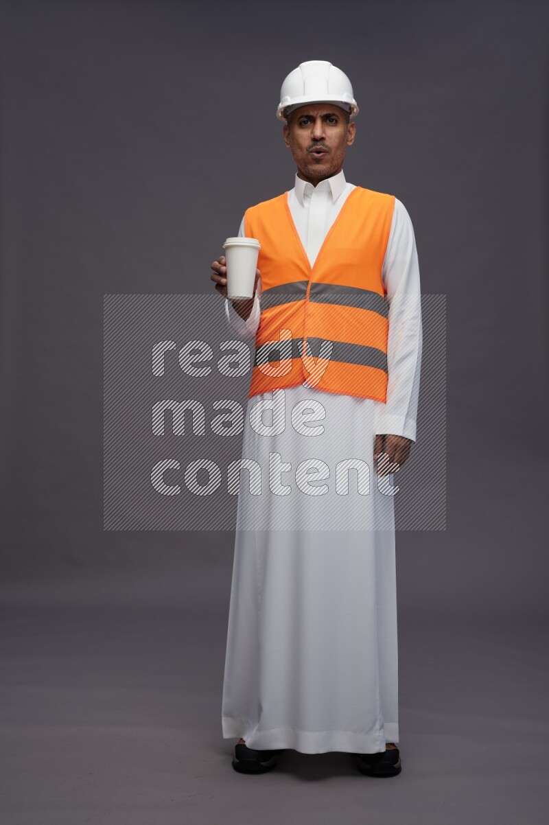 Saudi man wearing thob with engineer vest standing holding paper cup on gray background