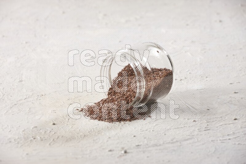 A glass jar full of flax seeds flipped and seeds spread out on a textured white flooring