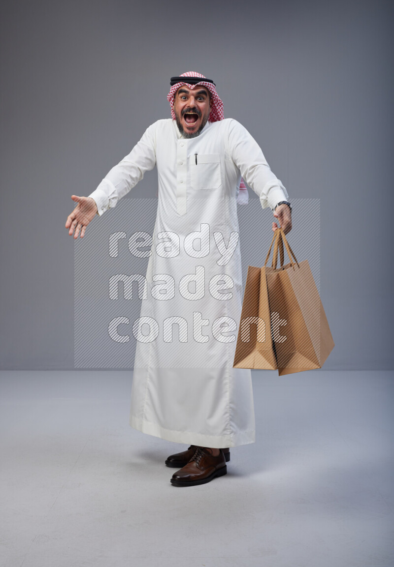 Saudi man Wearing Thob and red Shomag standing holding shopping bag on Gray background