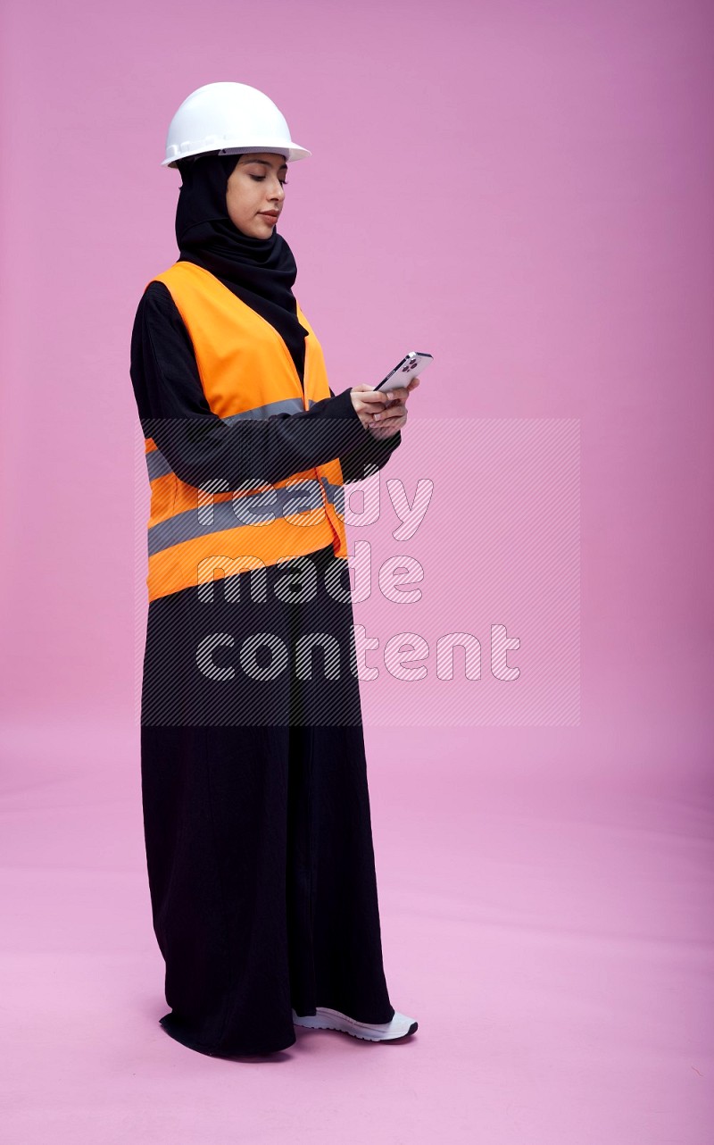 Saudi woman wearing Abaya with engineer vest and helmet standing texting on phone on pink background