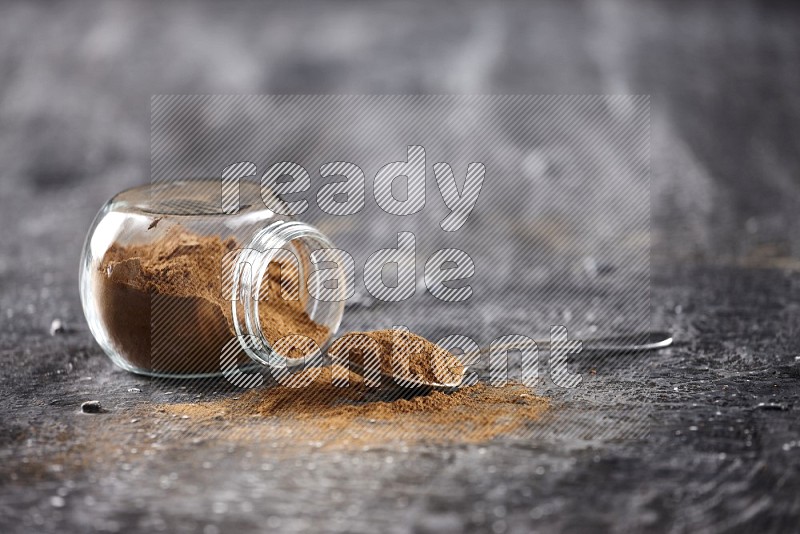 Herbal glass jar full of cinnamon powder flipped and a metal spoon on textured black background