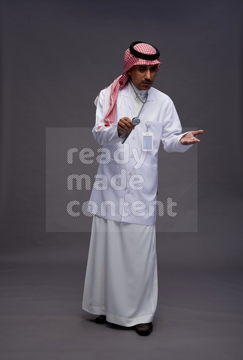 Saudi man wearing thob with lab coat and shomag with pocket employee badge with stethoscope standing interacting with the camera on gray background