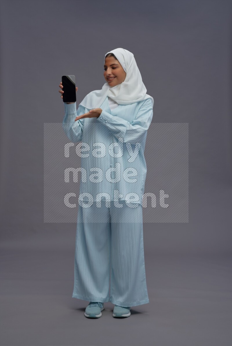 Saudi woman wearing hijab clothes standing showing phone to camera on gray background