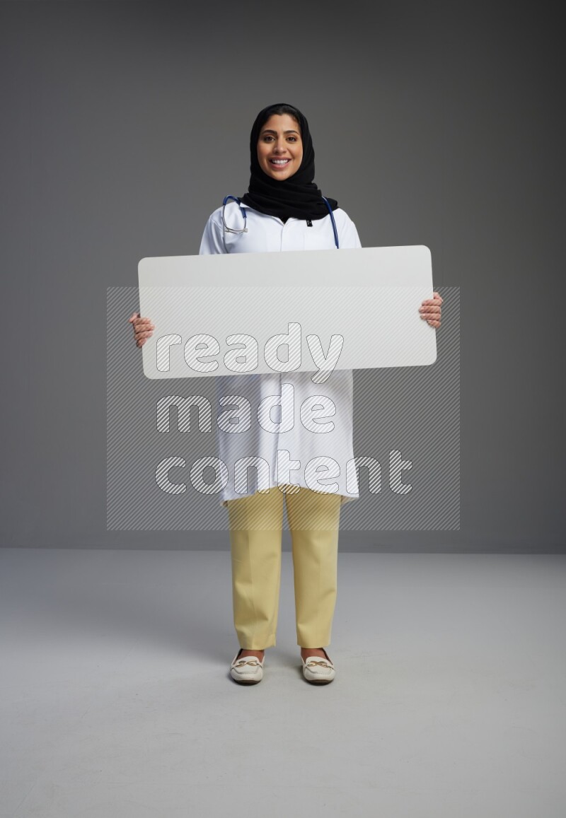 Saudi woman wearing lab coat with stethoscope standing holding board on Gray background