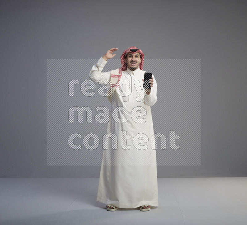 A Saudi man standing wearing thob and red shomag showing phone to camera on gray background