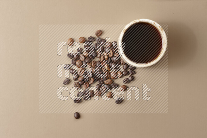 A beige pottery cup of coffee surrounded by roasted coffee beans on beige background