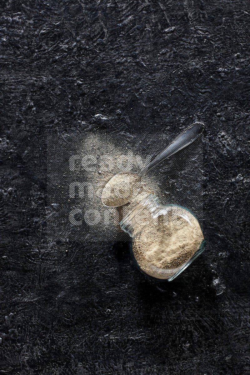 A flipped herbal glass jar and a metal spoon full of white pepper powder with spilled powder on textured black flooring