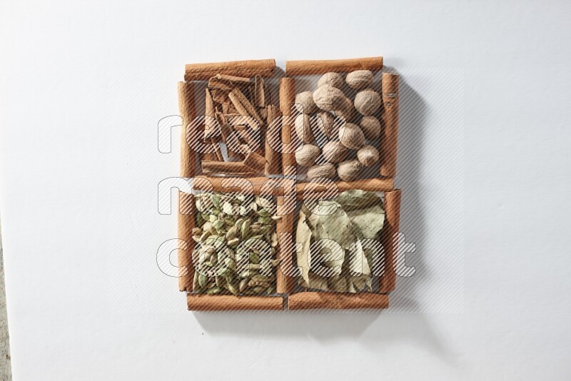 4 مربعات من أعواد القرفة ممتلئة بجوزة الطيب وأوراق الغار وحبوب الهيل والقرفة على أرضية بيضاء