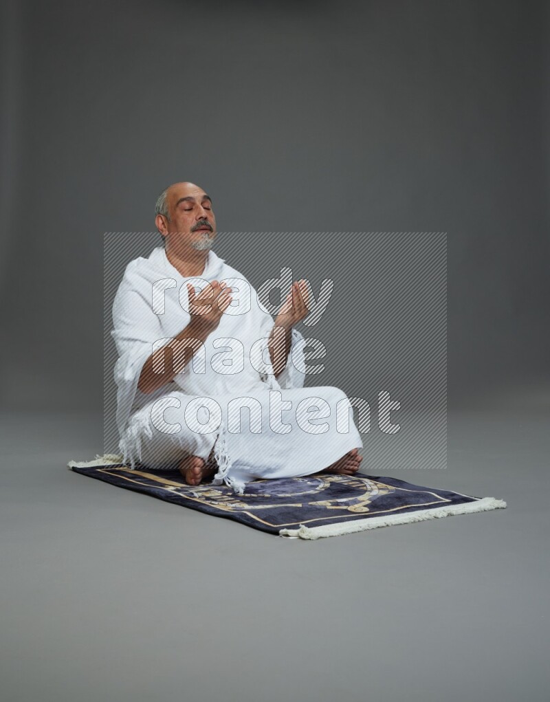 A man wearing Ehram sitting on prayer mat dua'a on gray background