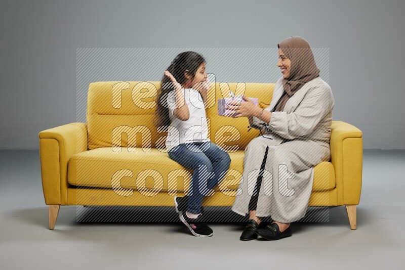 A girl sitting on a yellow sofa and giving a gift to her mother on gray background
