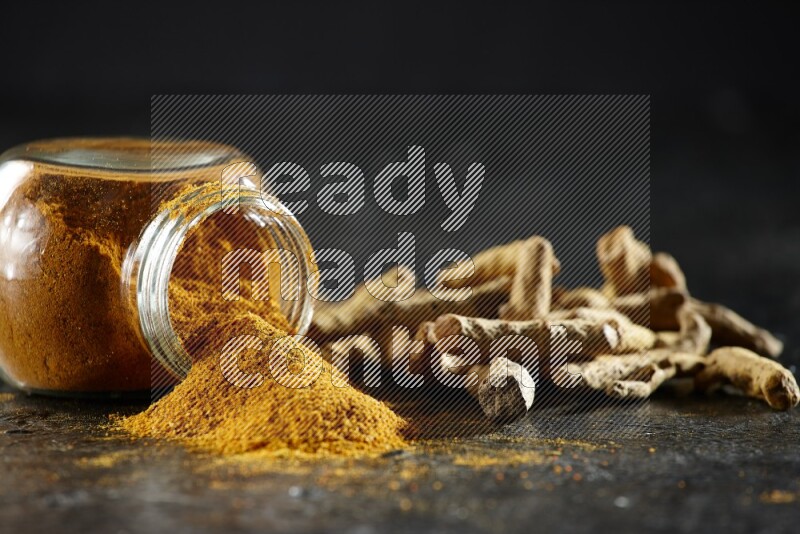 وعاء زجاجي للتوابل ممتلئ ببودرة الكركم مقلوب والبودرة تناثرت منه مع أصابع الكركم المجففة على خلفية سوداء