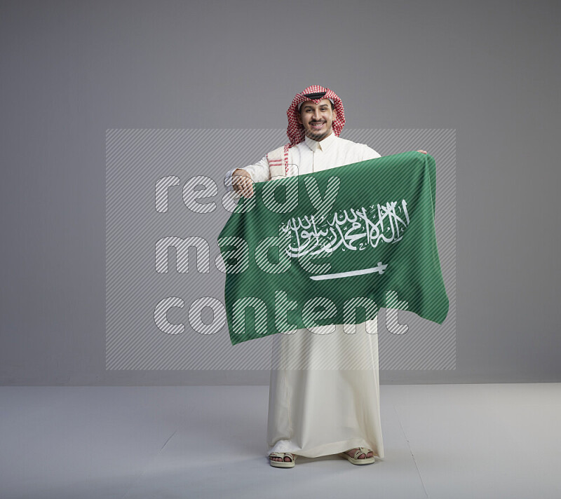 A saudi man standing wearing thob and red shomag holding big saudi flag on gray background