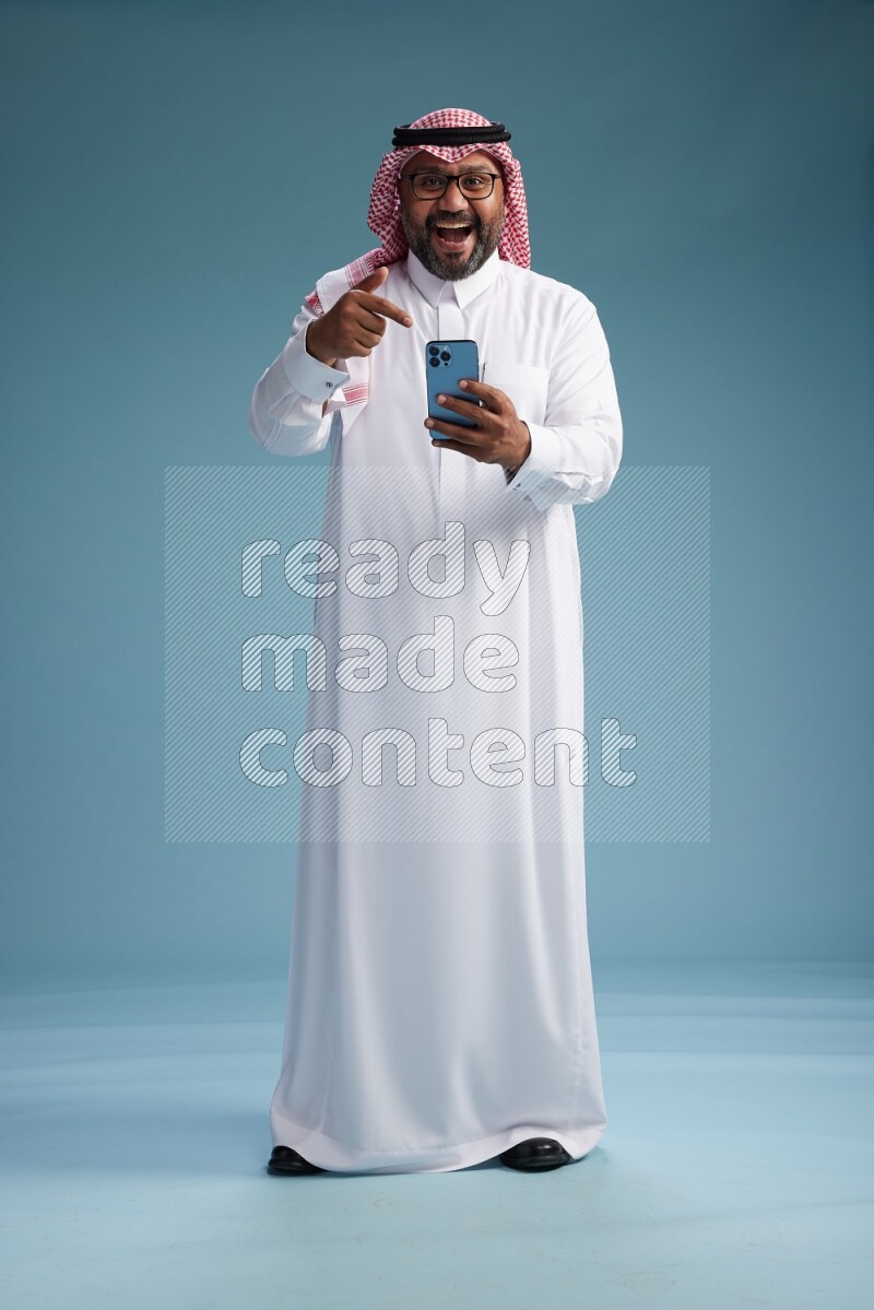 Saudi Man with shimag Standing texting on phone on blue background