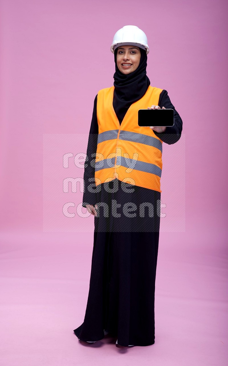 Saudi woman wearing Abaya with engineer vest and helmet standing showing phone to camera on pink background