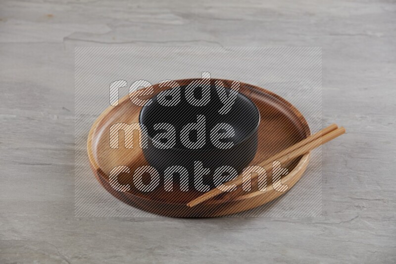 black ceramic round bowl on top of brown wood round plate and wood chopsticks, on grey textured countertop