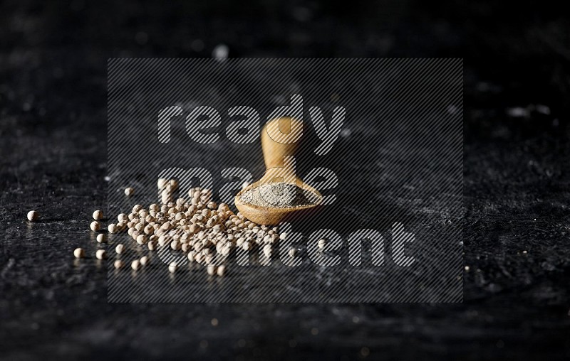 A wooden spoon full of white pepper powder with white pepper beads on textured black flooring
