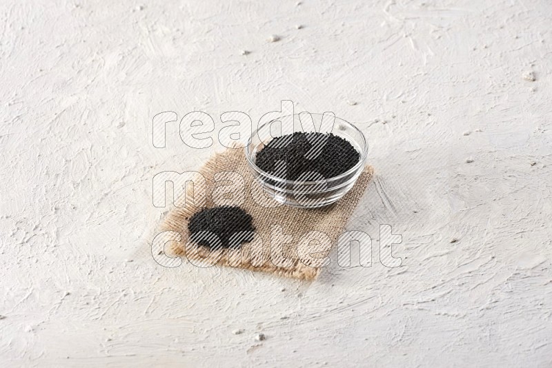A glass bowl full of black seeds on a burlap piece on textured white flooring