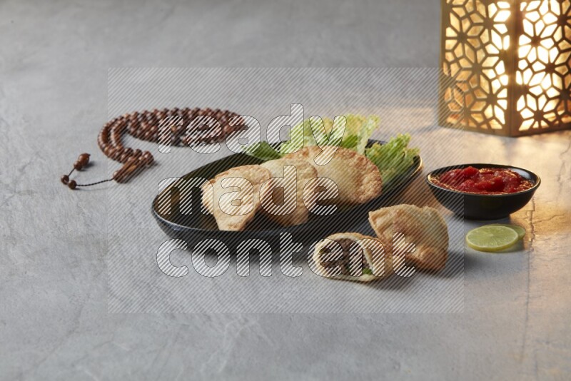 Three fried sambosas in an oval shaped black plate and a red sauce in a black round ramekin with a brown misbaha and a golden lantern on the side on a gray background
