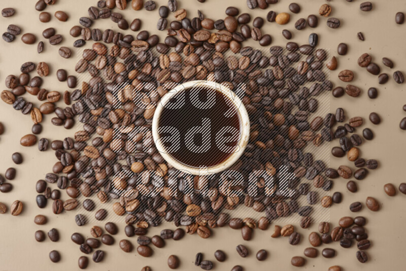 A beige pottery cup of coffee surrounded by roasted coffee beans on beige background