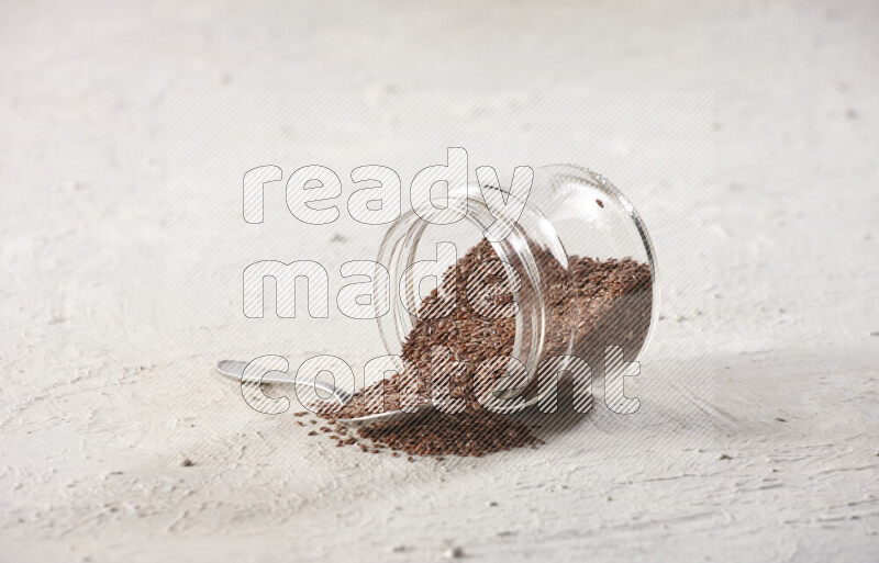 A glass spice jar full of flax seeds flipped with a metal spoon full of the seeds on a textured white flooring
