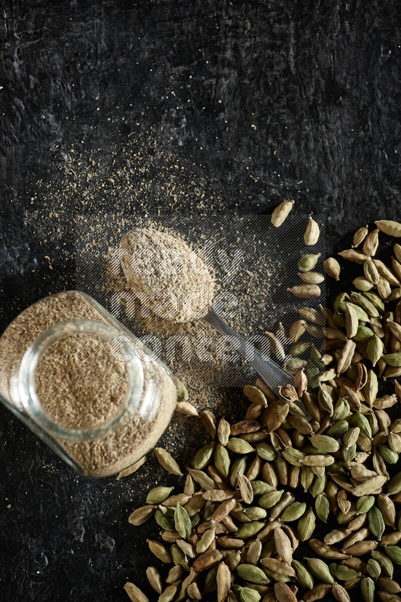 A glass spice jar and a metal spoon full of cardamom powder and cardamom seeds spreaded on textured black flooring
