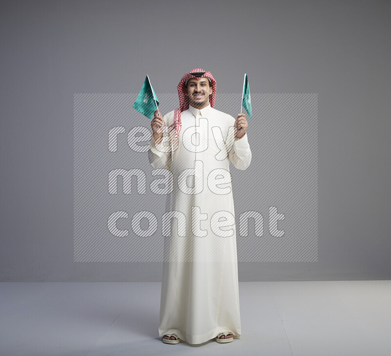 A Saudi man standing wearing thob and red shomag raising small Saudi flag on gray background