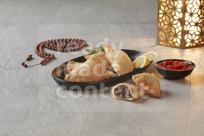 Four fried sambosas in an oval shaped black plate, beside a cut cheese sambosa, a brown misbaha and a golden lantern on a gray background