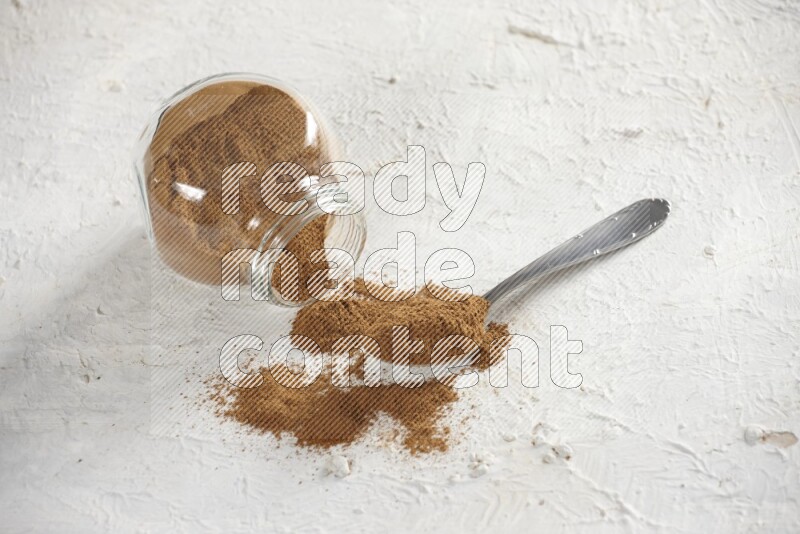 Flipped herbs glass jar full of cinnamon powder with a metal spoon full of powder on a textured white background