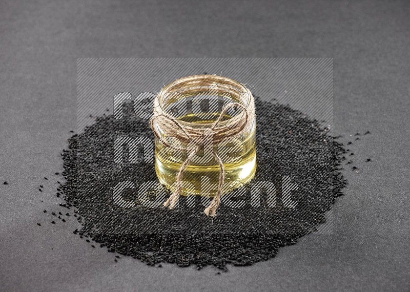 A glass jar full of black seeds oil surrounded by the seeds on a black flooring