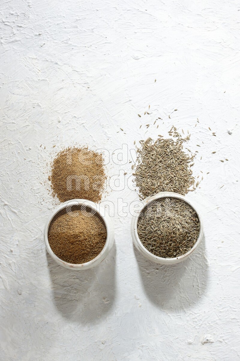 2 beige bowls full of cumin seeds and powder with spilled powder and seeds on textured white flooring