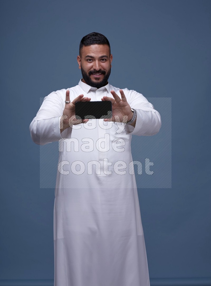 A man Showing his phone in Blue Background wearing Saudi Thob