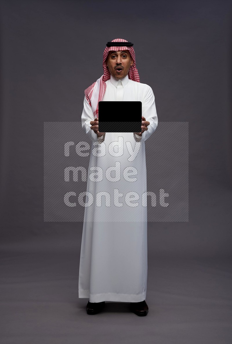 Saudi man wearing thob and shomag standing showing tablet to camera on gray background
